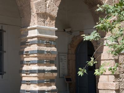 A close-up view of a stone arcade with a reinforced pillar featuring metal straps, leading to an arched doorway with a blue door and an informational sign.