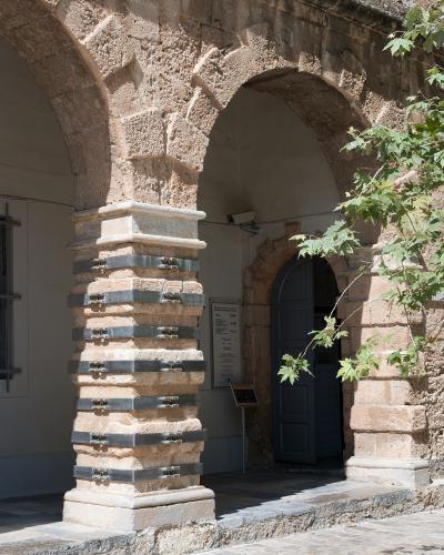 A close-up view of a stone arcade with a reinforced pillar featuring metal straps, leading to an arched doorway with a blue door and an informational sign.