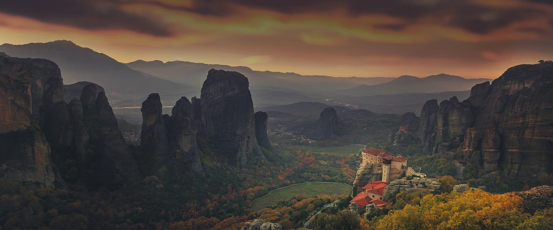 A wide landscape photograph showing a monastery with red-tiled roofs perched atop a massive sandstone rock pillar, surrounded by other towering rock formations and a valley with autumn foliage under a dramatic sunset sky.