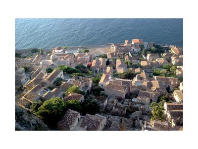 A high-angle view of a stone fortress wall and tower overlooking the sea and a cluster of buildings with terracotta roofs below.