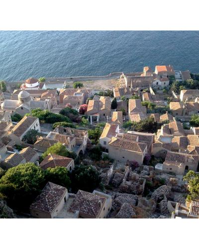 A high-angle view of a stone fortress wall and tower overlooking the sea and a cluster of buildings with terracotta roofs below.