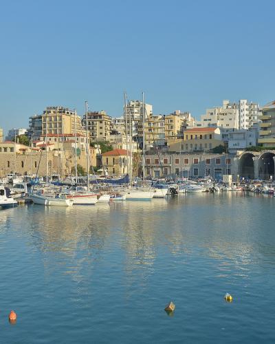 A view of the Venetian harbor in Heraklion, Crete, featuring the historic Venetian Arsenals (large stone buildings with arched openings) along the waterfront, with numerous boats moored in the harbor and modern buildings rising behind them.