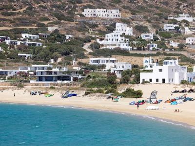 A sandy beach with turquoise water in the foreground, numerous windsurfing kites and gear on the sand, and a hillside covered in white modern villas and vegetation in the background.