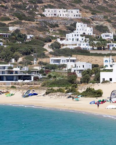 A sandy beach with turquoise water in the foreground, numerous windsurfing kites and gear on the sand, and a hillside covered in white modern villas and vegetation in the background.