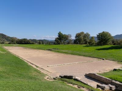 A long, rectangular ancient running track (stadion) with a sandy surface, stone starting blocks, and grassy surroundings under a blue sky.