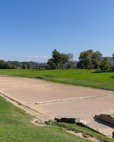 A long, rectangular ancient running track (stadion) with a sandy surface, stone starting blocks, and grassy surroundings under a blue sky.