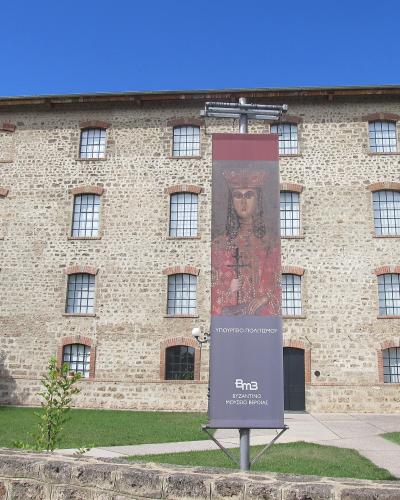 A large, three-story stone building with a grid of arched windows, featuring a vertical banner in the foreground displaying a Byzantine icon and Greek text.