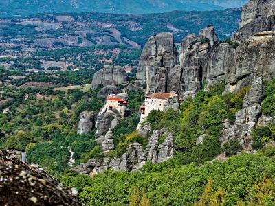 The image shows a wide landscape view of the Meteora rock formations in Greece, featuring two prominent monasteries (the Monastery of the Holy Trinity and the Monastery of Roussanou) situated on high rock pillars, with the vast Thessalian plain visib