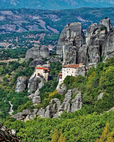 The image shows a wide landscape view of the Meteora rock formations in Greece, featuring two prominent monasteries (the Monastery of the Holy Trinity and the Monastery of Roussanou) situated on high rock pillars, with the vast Thessalian plain visib