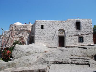 A whitewashed stone building with a domed roof and scaffolding on the left, situated on a rocky outcrop under a clear blue sky.