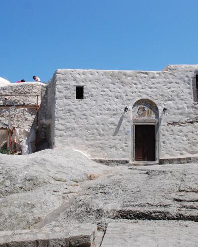 A whitewashed stone building with a domed roof and scaffolding on the left, situated on a rocky outcrop under a clear blue sky.