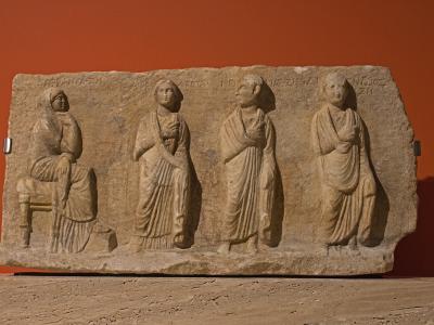 A stone funerary relief depicting four figures in classical dress against a solid terracotta-colored wall, with Greek inscriptions visible along the top edge.