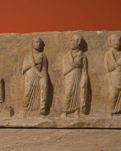 A stone funerary relief depicting four figures in classical dress against a solid terracotta-colored wall, with Greek inscriptions visible along the top edge.