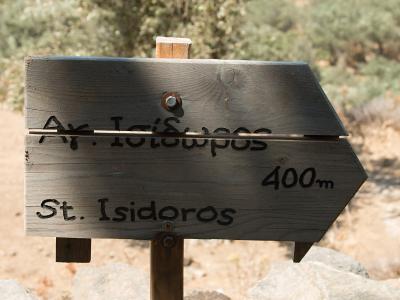 A wooden directional signpost with Greek text "Αγ. Ισίδωρος" and English text "St. Isidoros" indicating a distance of 400m, set against a dry, scrubby landscape.