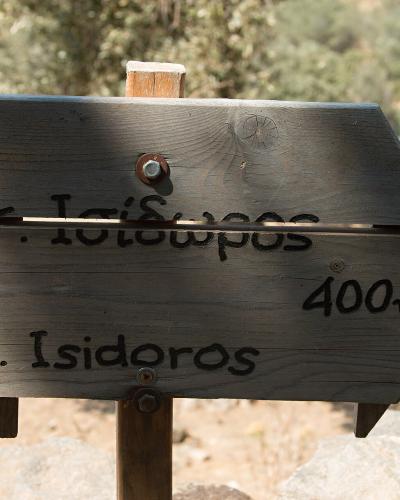A wooden directional signpost with Greek text "Αγ. Ισίδωρος" and English text "St. Isidoros" indicating a distance of 400m, set against a dry, scrubby landscape.