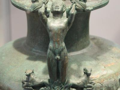 A close-up photograph of the lid and upper neck of an ancient bronze hydria, featuring a central nude male figure supporting the lid rim, flanked by sphinxes and resting on a base with two recumbent animals.