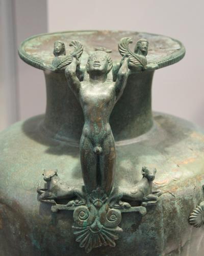 A close-up photograph of the lid and upper neck of an ancient bronze hydria, featuring a central nude male figure supporting the lid rim, flanked by sphinxes and resting on a base with two recumbent animals.