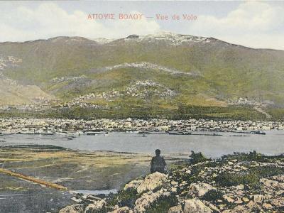 A vintage colorized postcard showing a coastal city and harbor nestled at the foot of a large mountain, with a figure sitting on a rocky outcrop in the foreground.
