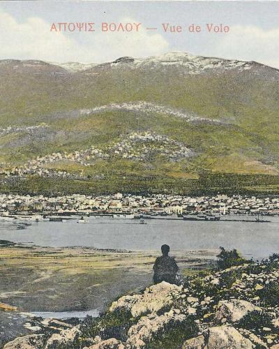 A vintage colorized postcard showing a coastal city and harbor nestled at the foot of a large mountain, with a figure sitting on a rocky outcrop in the foreground.