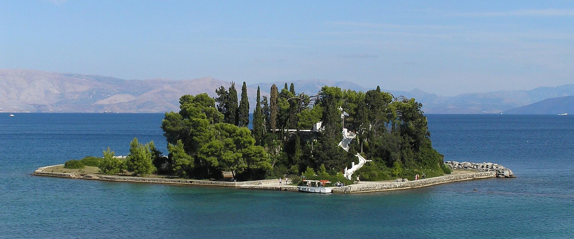 A small, tree-covered islet featuring a prominent white church and a stone causeway, set against a backdrop of blue sea and distant mountains.