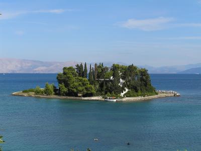 A small, tree-covered islet featuring a prominent white church and a stone causeway, set against a backdrop of blue sea and distant mountains.