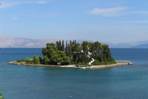 A small, tree-covered islet featuring a prominent white church and a stone causeway, set against a backdrop of blue sea and distant mountains.