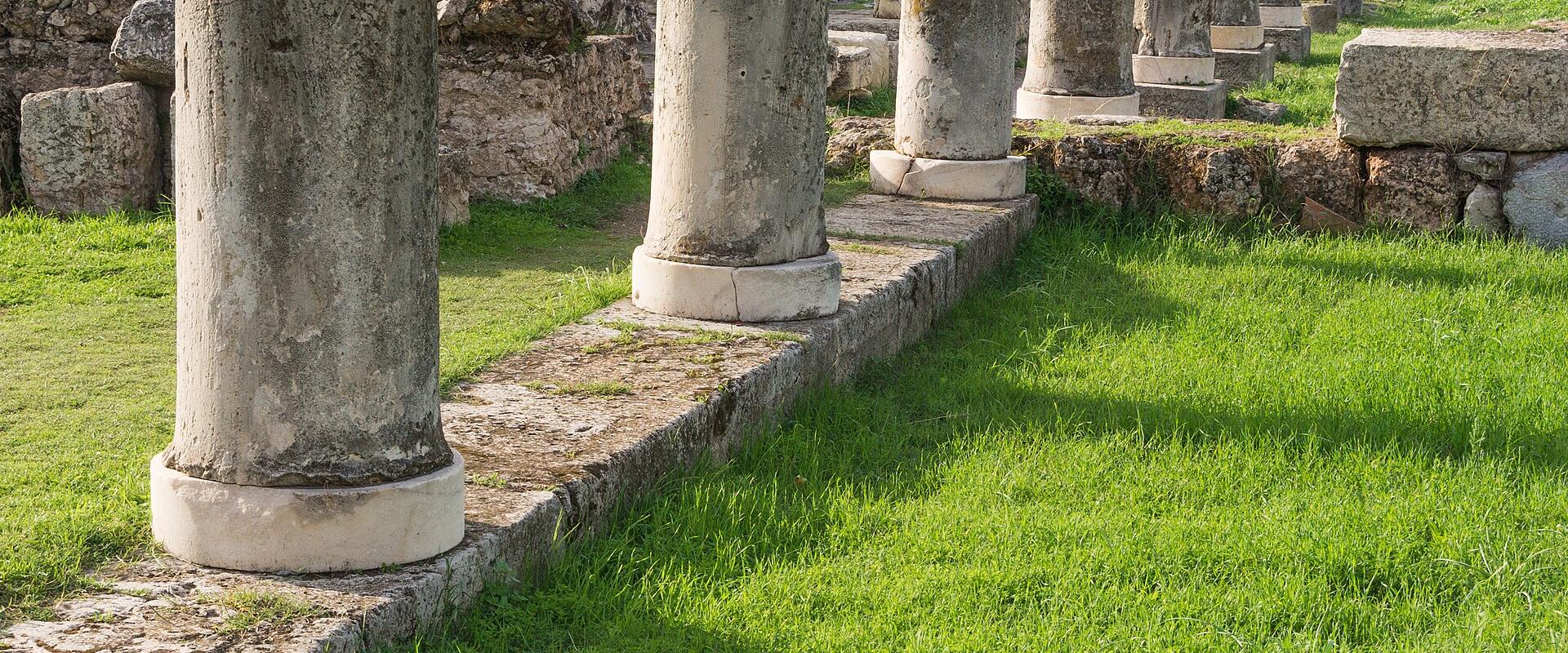 A row of weathered stone column drums and bases set in a grassy field with ancient stone walls and ruins in the background.