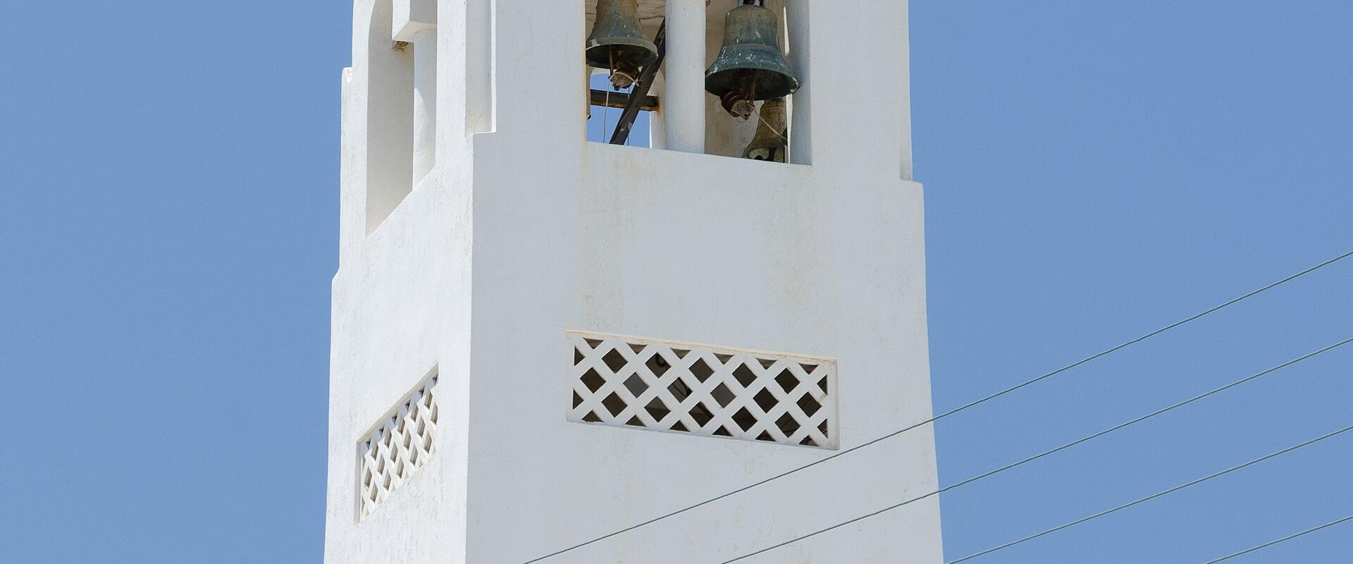 A close-up view of a white-washed bell tower with arched openings containing bells, topped with a cross, set against a clear blue sky.