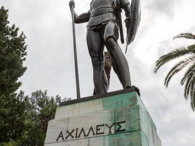A low-angle shot of a large bronze statue of a warrior holding a spear and shield, standing on a marble and copper-clad pedestal inscribed with the Greek word "ΑΧΙΛΛΕΥΣ" (Achilles).
