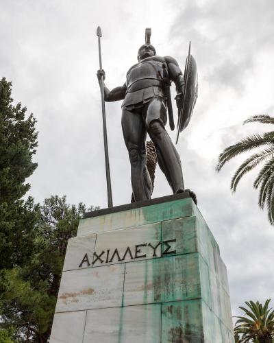 A low-angle shot of a large bronze statue of a warrior holding a spear and shield, standing on a marble and copper-clad pedestal inscribed with the Greek word "ΑΧΙΛΛΕΥΣ" (Achilles).