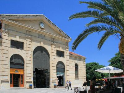 A large neoclassical building with a central pediment, a clock, and arched entrances, situated in a paved public square with a palm tree and market stalls nearby.