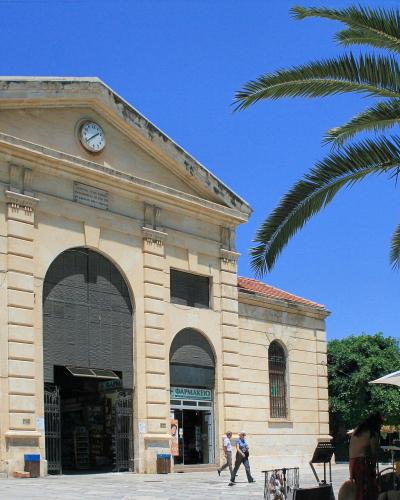 A large neoclassical building with a central pediment, a clock, and arched entrances, situated in a paved public square with a palm tree and market stalls nearby.