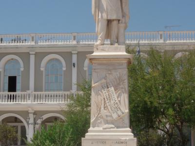 A marble statue of a standing man on a tall pedestal with Greek inscriptions, set against a large neoclassical building with arched windows and a balustrade.