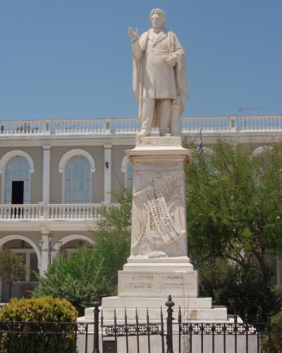 A marble statue of a standing man on a tall pedestal with Greek inscriptions, set against a large neoclassical building with arched windows and a balustrade.