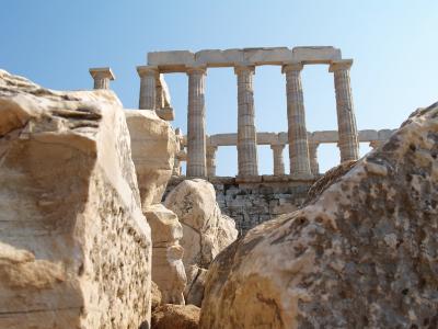 A low-angle view of ancient Greek Doric columns and stone ruins set against a clear blue sky, framed by large rocks in the foreground.