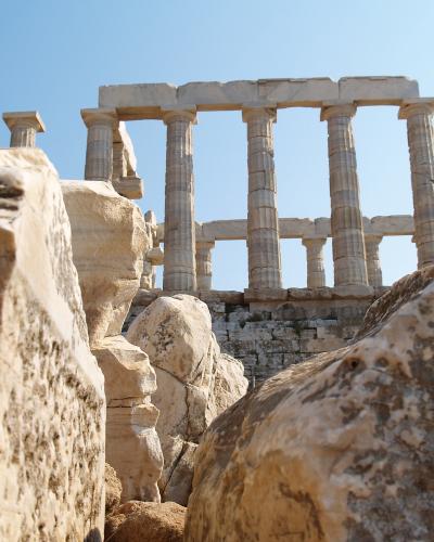 A low-angle view of ancient Greek Doric columns and stone ruins set against a clear blue sky, framed by large rocks in the foreground.