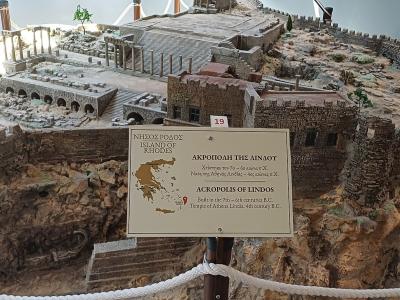 A detailed architectural scale model of an ancient fortified settlement on a rocky hill, displayed indoors behind a rope barrier with an informational sign in the foreground.