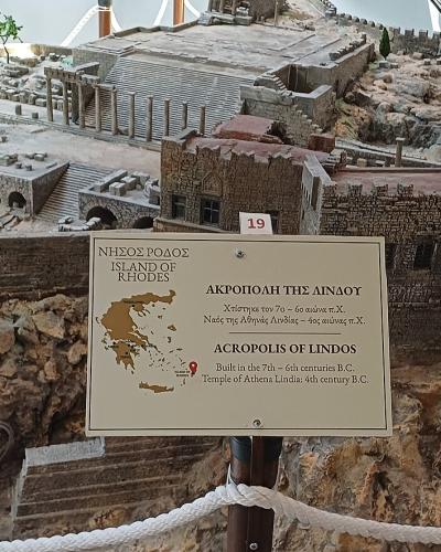 A detailed architectural scale model of an ancient fortified settlement on a rocky hill, displayed indoors behind a rope barrier with an informational sign in the foreground.