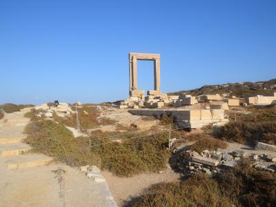A large, freestanding marble doorway (Portara) standing amidst ancient stone ruins and dry vegetation under a clear blue sky.