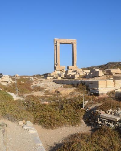 A large, freestanding marble doorway (Portara) standing amidst ancient stone ruins and dry vegetation under a clear blue sky.