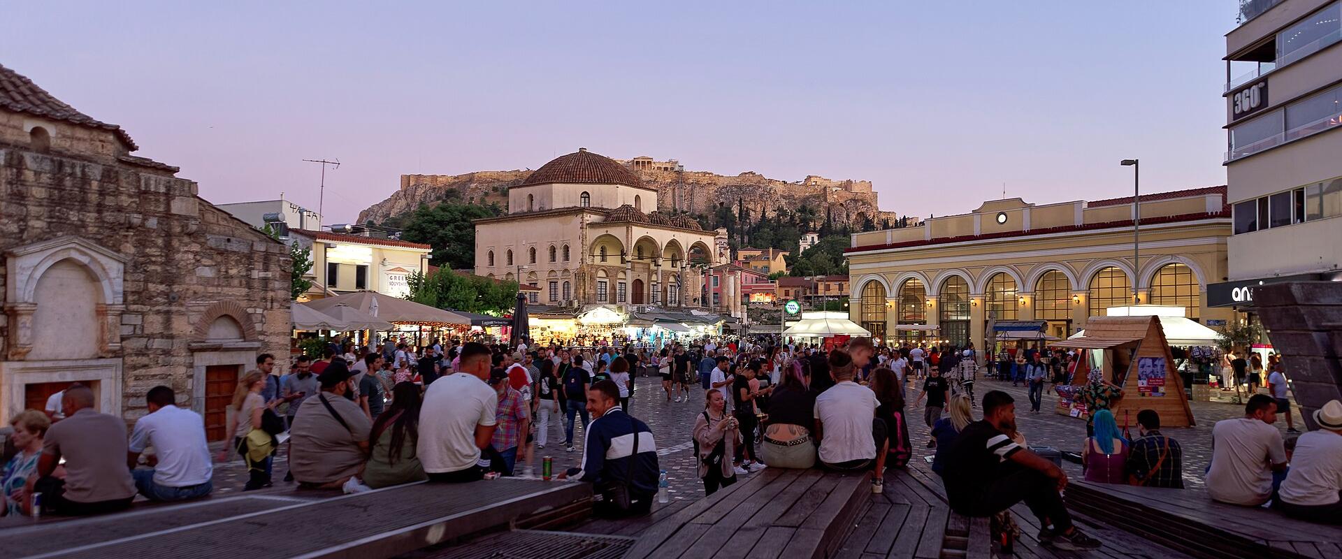 A crowded public square at dusk featuring a large Ottoman-era mosque with a dome and minaret in the center, the Acropolis hill with the Parthenon visible in the background, and a long wooden seating structure in the foreground.