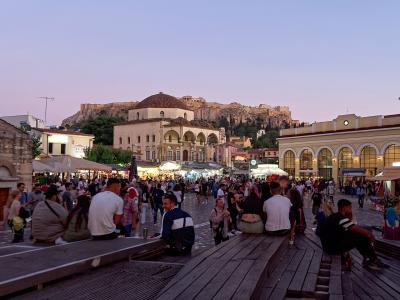 A crowded public square at dusk featuring a large Ottoman-era mosque with a dome and minaret in the center, the Acropolis hill with the Parthenon visible in the background, and a long wooden seating structure in the foreground.