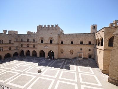 A high-angle view of a large, sunlit stone courtyard surrounded by medieval buildings with arched arcades and crenellated towers, featuring a distinctive geometric pavement pattern.