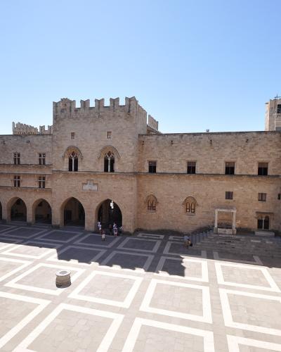 A high-angle view of a large, sunlit stone courtyard surrounded by medieval buildings with arched arcades and crenellated towers, featuring a distinctive geometric pavement pattern.