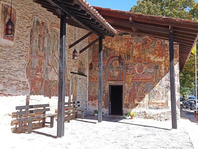 A stone building with extensive exterior frescoes under a wooden porch roof, featuring wooden benches on a paved courtyard.