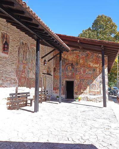 A stone building with extensive exterior frescoes under a wooden porch roof, featuring wooden benches on a paved courtyard.