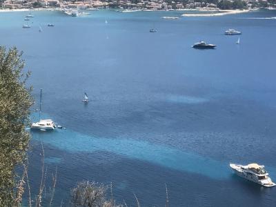A high-angle view of a coastal bay with numerous boats anchored in blue water, a small town along the shoreline, and forested hills in the background.