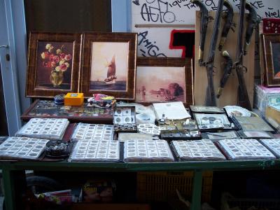 A street vendor's display table featuring framed paintings, old flintlock muskets, and numerous binders of coins and stamps.