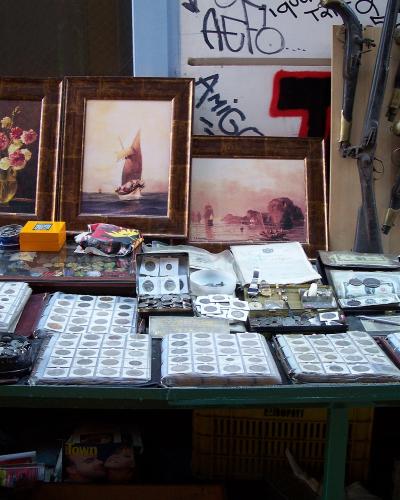 A street vendor's display table featuring framed paintings, old flintlock muskets, and numerous binders of coins and stamps.