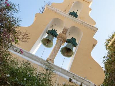 A low-angle shot of a yellow church bell tower with three bells, set against a blue sky and surrounded by green vegetation with white and pink flowers.